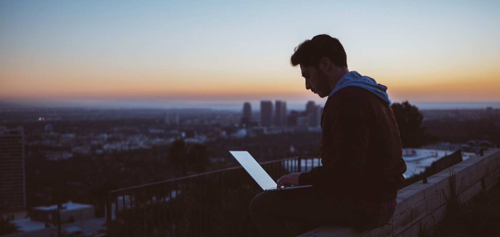 Male student sitting elevated on a wall with a laptop, in the background a large city.