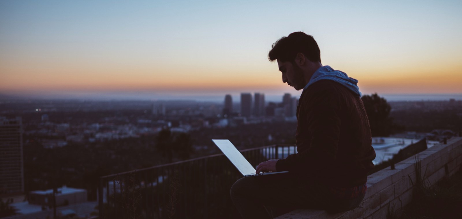 avi-richards-Z3ownETsdNQ-unsplash(2).jpg Male student sitting elevated on a wall with a laptop, in the background a large city.